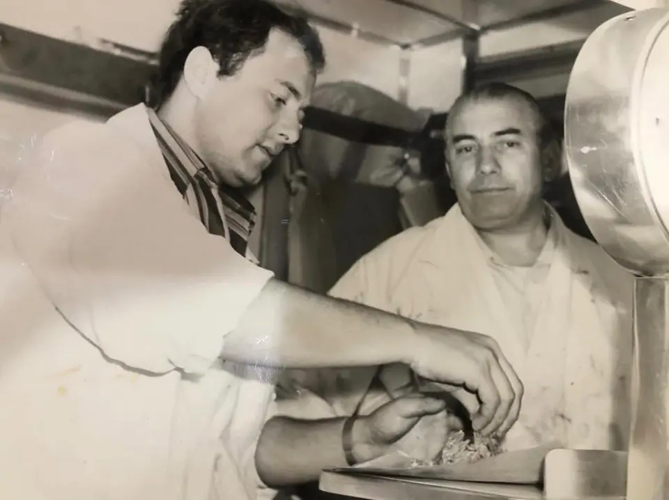 Foto d’epoca in bianco e nero: due macellai in camice bianco al banco, uno prepara la carne accanto alla bilancia